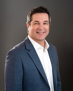 Man in a blue suit jacket and white shirt smiles at the camera against a plain, dark gray background.