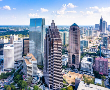 Aerial view of a city skyline with modern skyscrapers, glass buildings, and green spaces under a blue sky with scattered clouds.