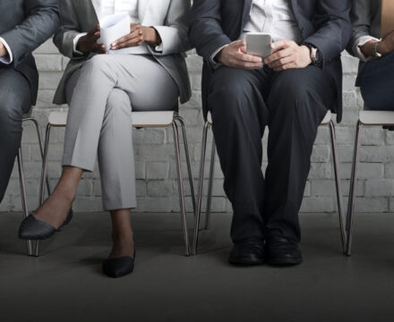 Four people in business attire sit on chairs against a white brick wall, their faces out of frame. Each is holding a mobile device, notebook, or paper, suggesting they are waiting for an interview or meeting.
