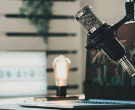 A podcasting setup with a microphone, a laptop, a glowing lightbulb, and an illuminated ON AIR sign on a desk.