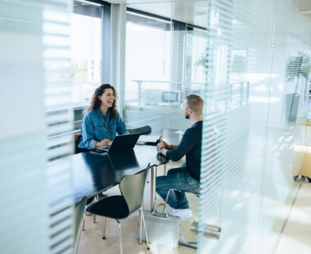 Two people sit across from each other at a table in a modern office, talking and smiling. One person uses a laptop, while the other listens. The room has large windows and glass walls, letting in natural light.