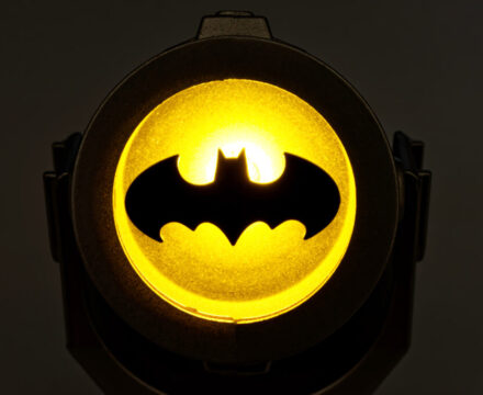 A glowing yellow Bat-Signal projector shines the iconic black bat symbol against a dark background.