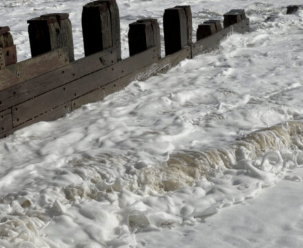 Waves crash against a weathered wooden seawall, creating thick sea foam that covers the water and shoreline.
