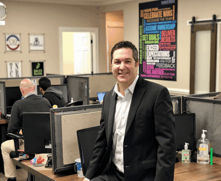 A man in a suit smiles while sitting on a desk in a modern office with several people working at computers in the background. Motivational posters and hand sanitizer are visible on and around the desks.