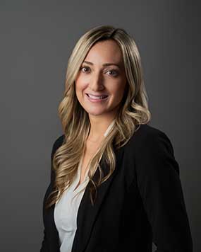 Smiling woman with long, wavy blonde hair, wearing a black blazer over a white top, poses against a plain gray background.