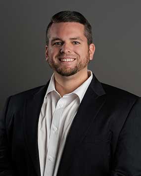 Man with short dark hair and beard, wearing a black suit jacket over a white dress shirt, smiling, and standing in front of a plain gray background.